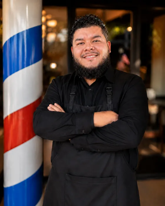 Ernesto Sedano, barber at Dandies, smiling beside the barber pole